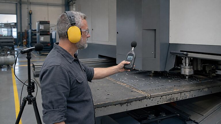 Un técnico de acústica con protectores auditivos y gafas de seguridad mide con un sonómetro el nivel de presión acústica en el puesto de mando de una punzonadora para documentar las emisiones sonoras.