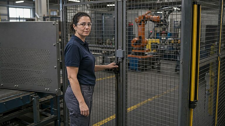 Una técnica de mantenimiento con gafas de seguridad apoya la mano en la puerta cerrada de una celda robotizada protegida por resguardo perimetral con interruptor de enclavamiento, un resguardo fijo atornillado a la izquierda y una cortina óptica a la derecha.